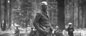 A uniformed middle-aged man standing with hands in pockets in a grove of trees