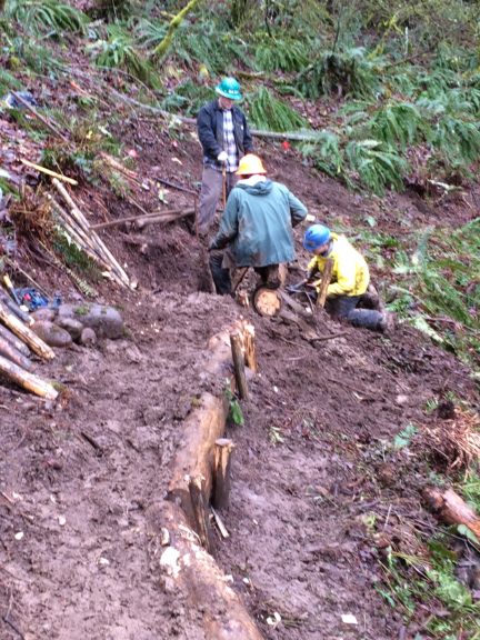 Three workers wearing hard hats are clustered at a point on a muddy trail as one wields a pair of loppers.