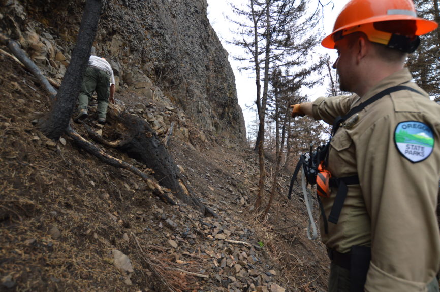 A park ranger points to a sloping rocky trail at the base of a cliff with burnt trees around.