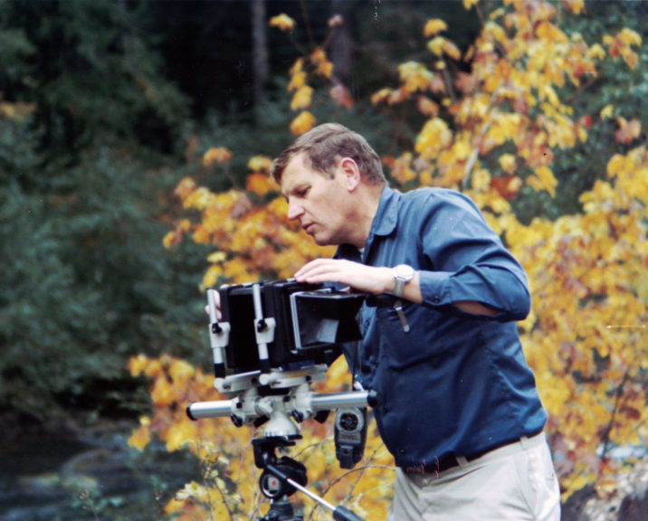 A man adjusts a large camera on a tripod with yellow fall foliage in the background. 