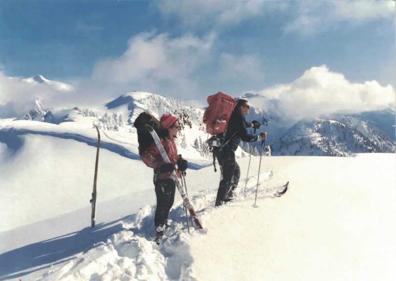 A man on skis and a woman who has taken off her skis stand in deep snow on a ridgeline and look out at a view of snowy mountains.