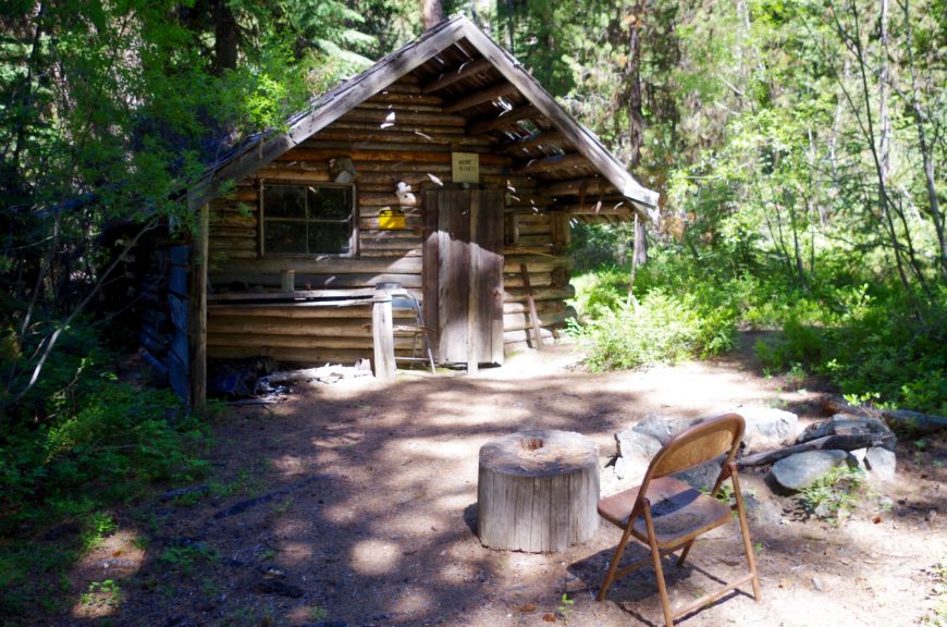 A small log cabin in a clearing with a rusted folding chair facing it.