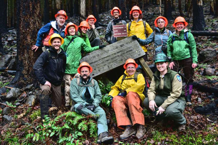 A dozen people in hard hats and rain gear cluster around a fallen over wood sign.
