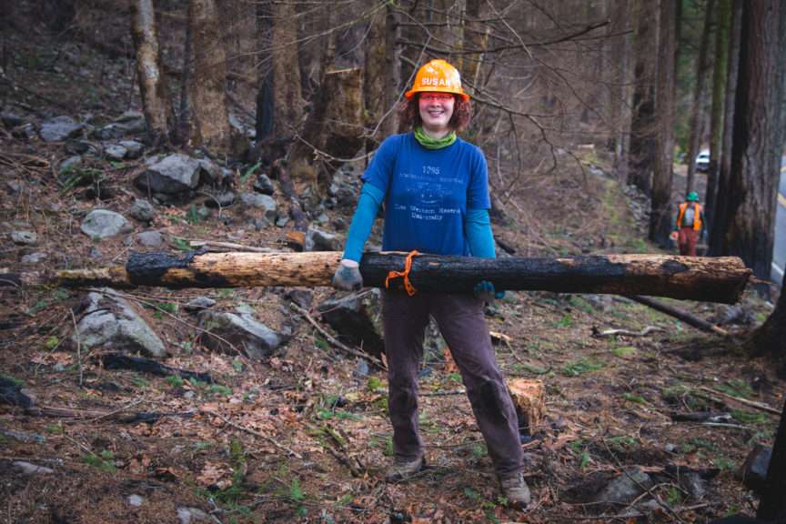 A woman wearing a hard hat carrying a burned log.