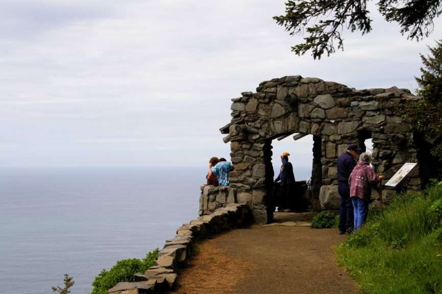 People standing in and in front of a small stone building on a cliff above an expanse of ocean.