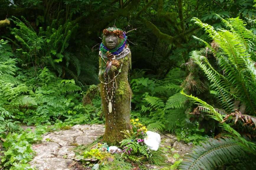 A stone statue draped in beads against a backdrop of moss, ferns, and conifers.