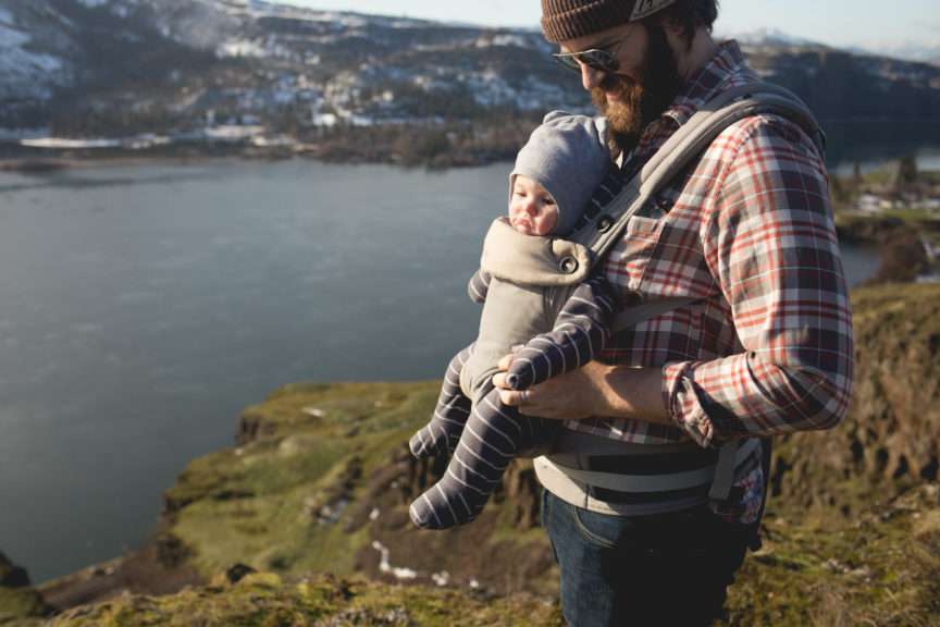 A man with a baby in a baby carrier on his chest standing on a cliff high above a wide river.
