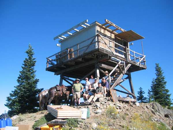A group of people sit and stand on the ground with construction materials in front of a cabin on stilts.