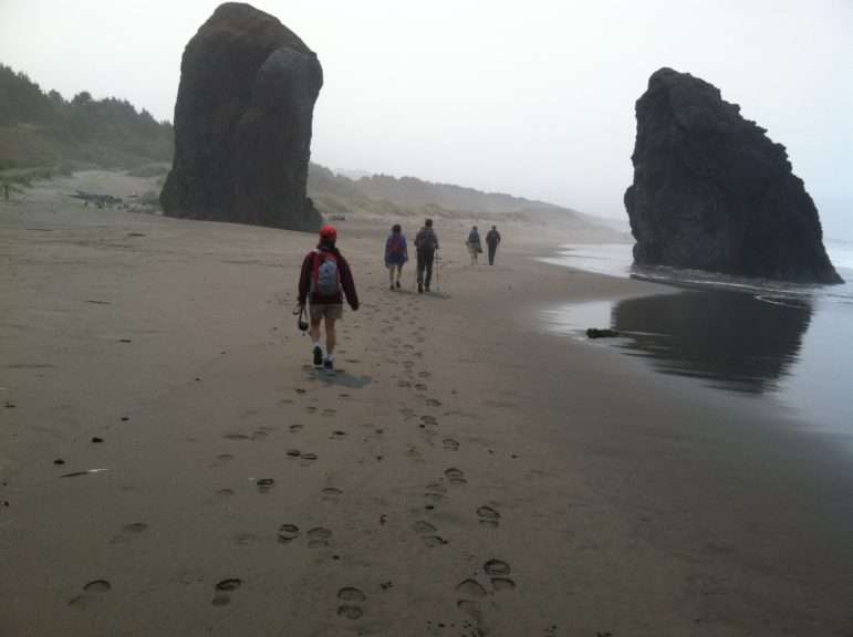 Five people walk along a beach between two large monoliths rising out of the sand.
