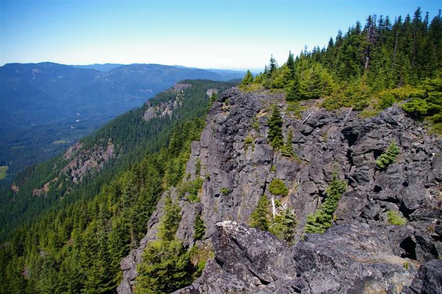 An exposed rock cliff topped by coniferous trees with mountains in the distance under a blue sky.