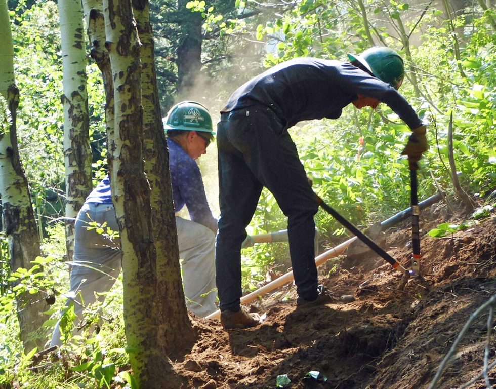 Two workers in hardhats apply tools to a trail on steep terrain.