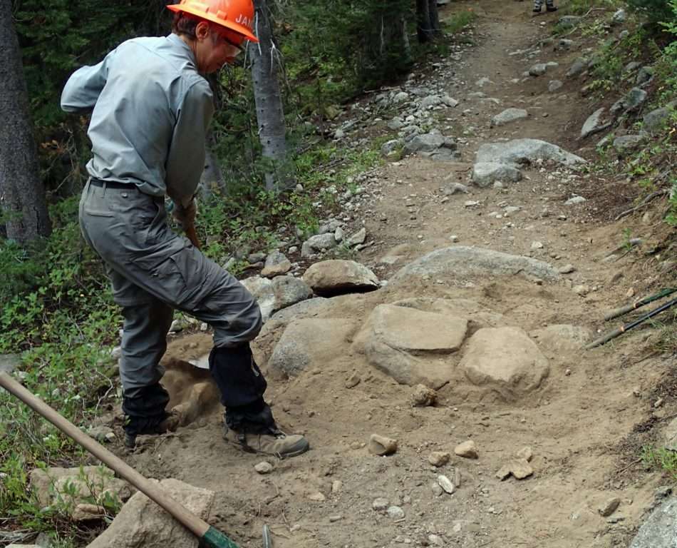 A hard-hatted man standing above several boulders and a small trench crossing a trail