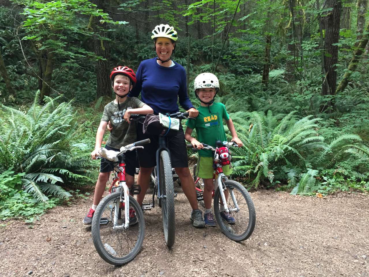 A woman flanked by a boy on either side, all smiling as they sit astride bicycles against a green backdrop of sword ferns and trees.