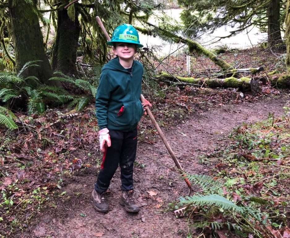 A young boy wearing a hardhat holds a rake on a trail.