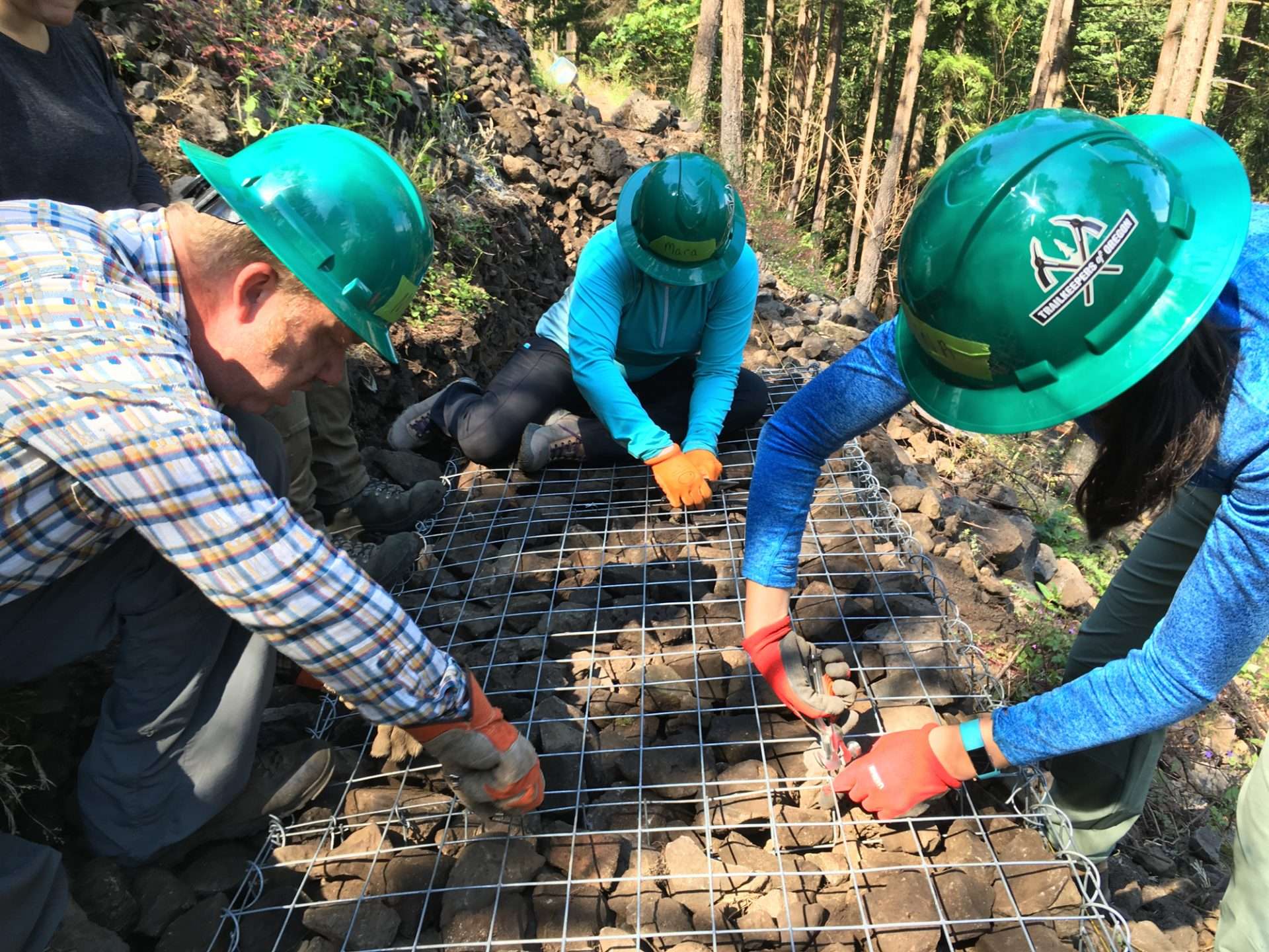 Three people in green hard hats leaning over a metal grid with rocks beneath it.