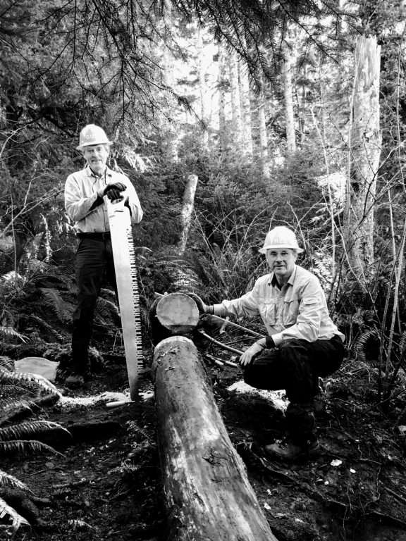 Two men on either side of a cut-through log, one standing with his hands on a long saw, the other kneeling by the cut log.