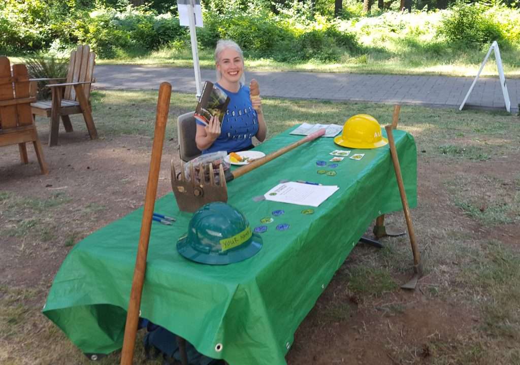 A smiling woman holding a book and an ice cream cone sits outdoors at a table which has hardhats and a McLeod trail tool on it.