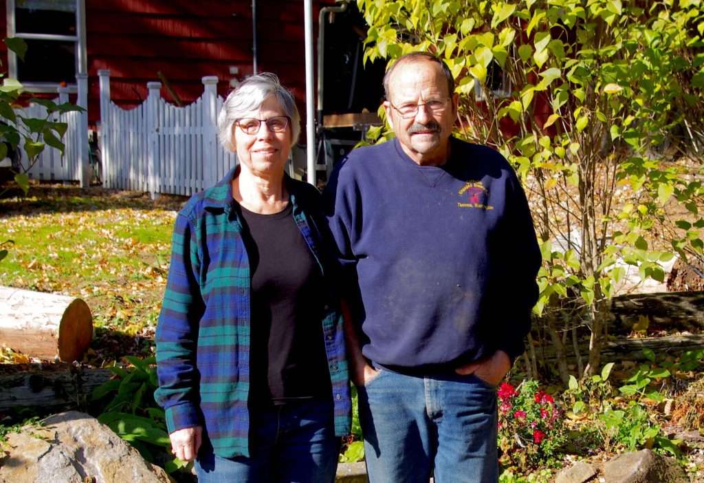 A man and a woman stand in autumnal sunshine in front of a red house.