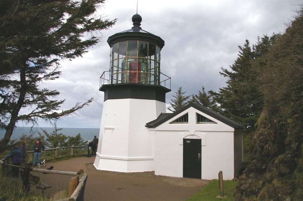 A squat black-and-white lighthouse with an adjoining building and ocean in background.