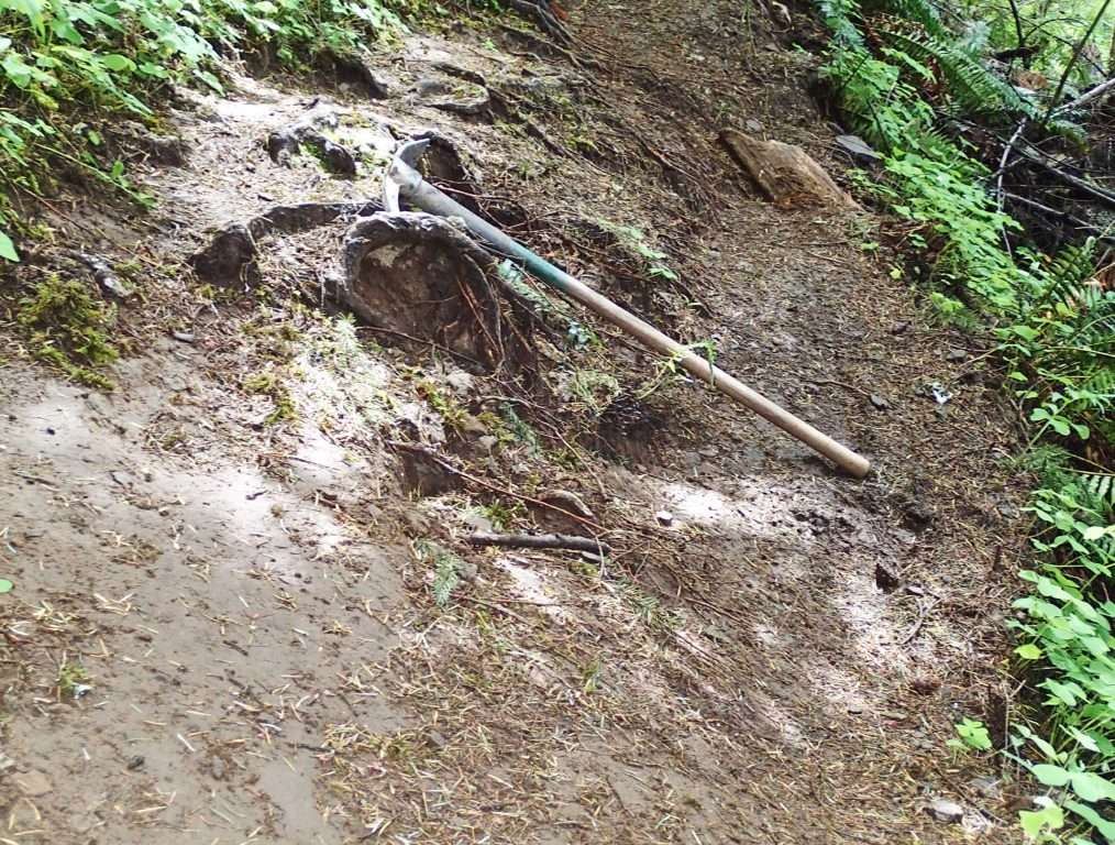 A trail tool (pick mattock) rests on a section of trail that has subsided down a slope.