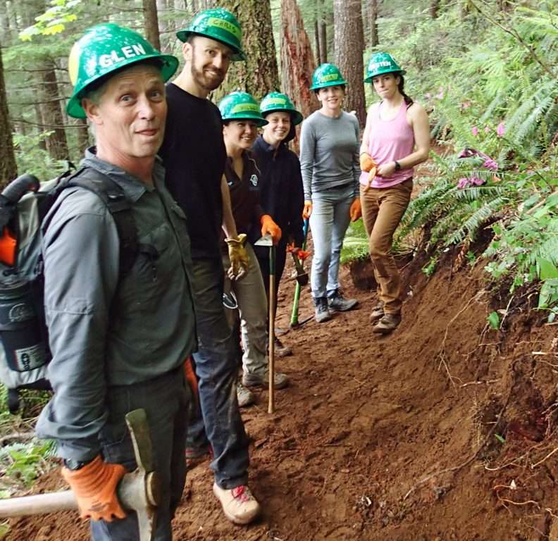 Six trail workers in green hard hats stand on a restored section of trail and smile at the camera.