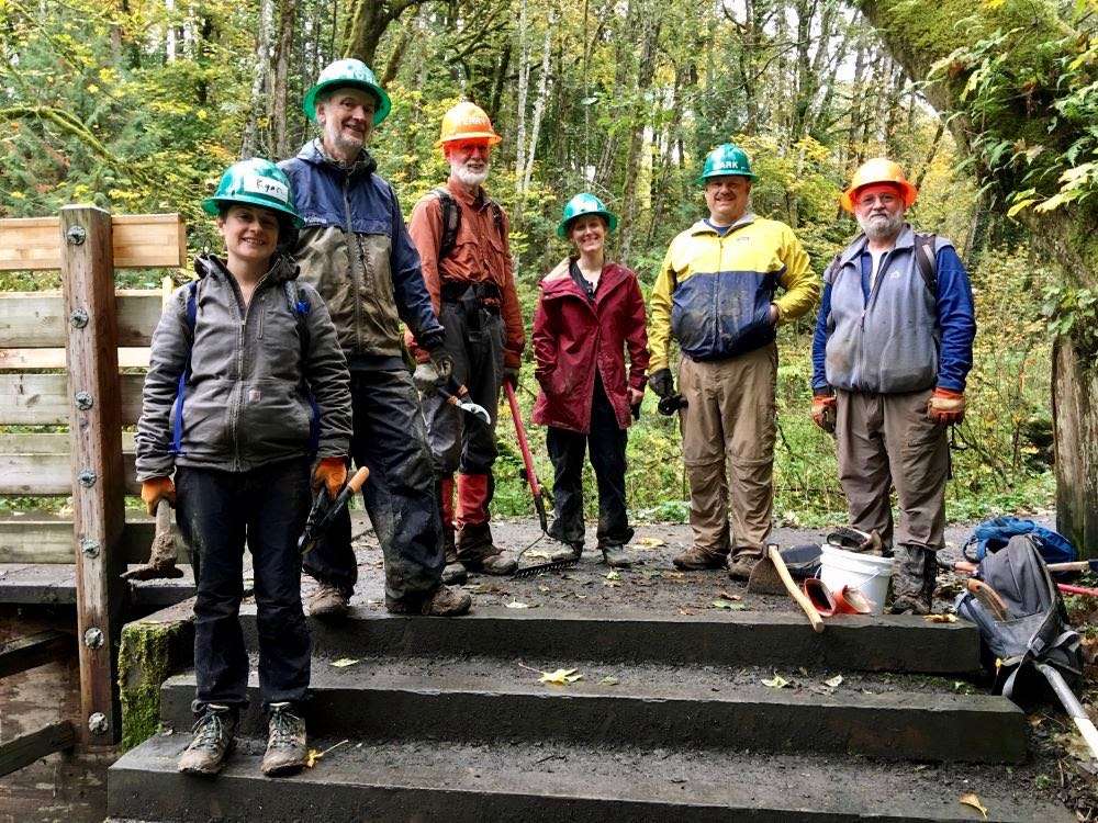 Six trail volunteers in hard hats posing above a short staircase on a trail in a wooded area.