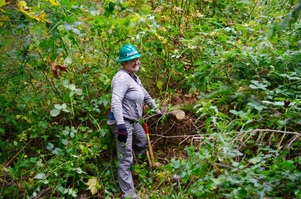 A trail volunteer in a green hard hat posing by a blackberry thicket.