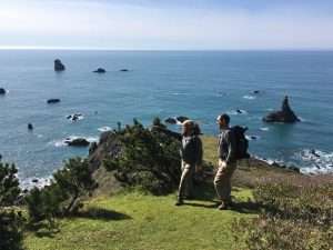 Two men walk a grassy coastal headland above a sparkling blue sea peppered with rock islands.