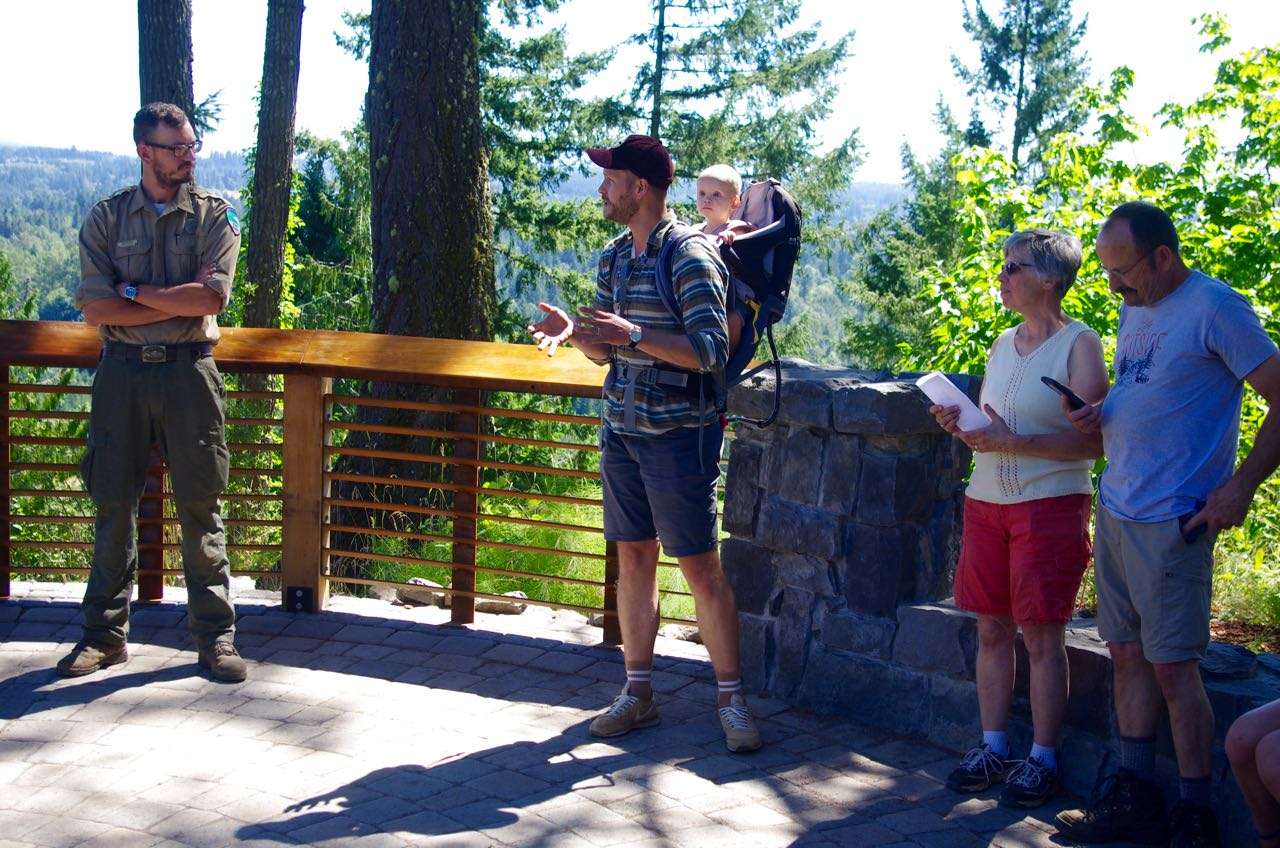 A park ranger in uniform, a bearded, gesturing man with a baby on his back, and another man and a woman stand with backs to a guard rail.