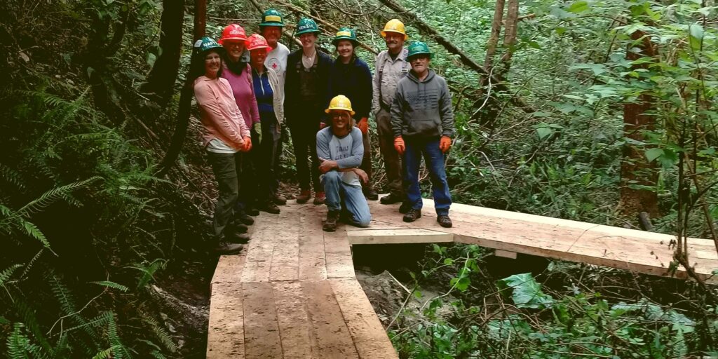 A group of people in hard hats stand smiling on a newly built boardwalk in the woods.