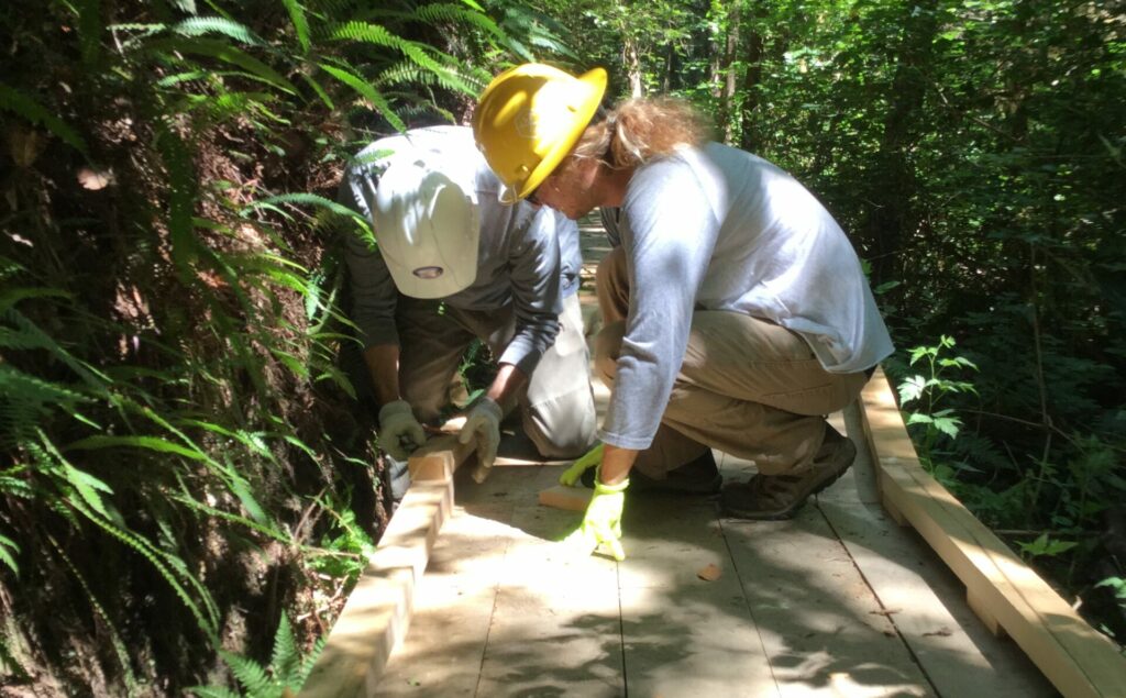 Two people in hard hats work on their knees to install a piece of decking on a bardwalk.