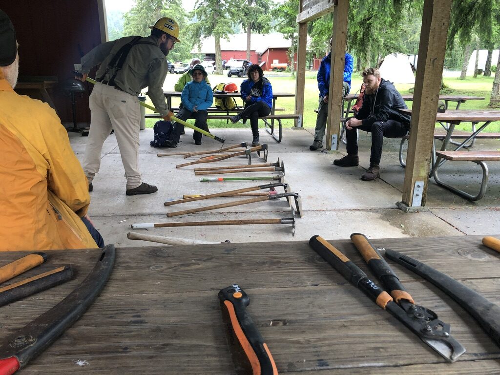 A variety of long handled hand tools are laid out under a picnic shelter. A person in hard hat demonstrates how to use one of the tools to a gather group