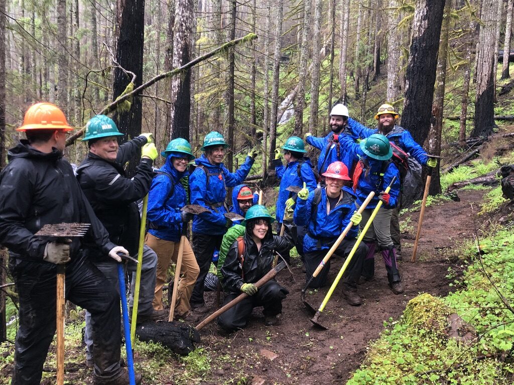A group of people in rain jackets and hard hats stand on a hiking trail in the woods, pointing at a waterfall behind them and through the trees