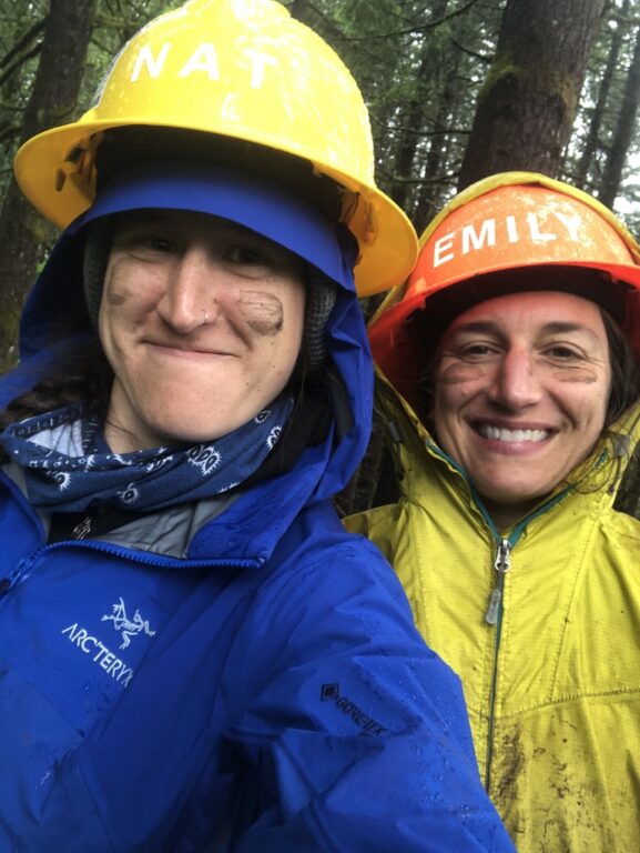 A close up photo of the faces of two people in rain gear and hard hats with muddy cheeks smiling at the camera