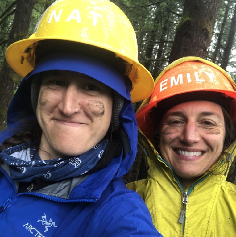 A close up of two faces. One person wears a blue jacket and a yellow hat, the other wears a yellow jacket and an orange hat. They both have dirt on their faces and are smiling. 