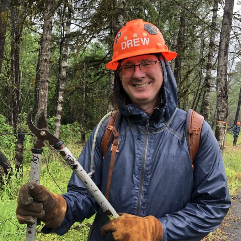 a person in a dark blue rain jacket wearing an orange hard hat holds a pair of pruning loppers and smiles at the camera. 