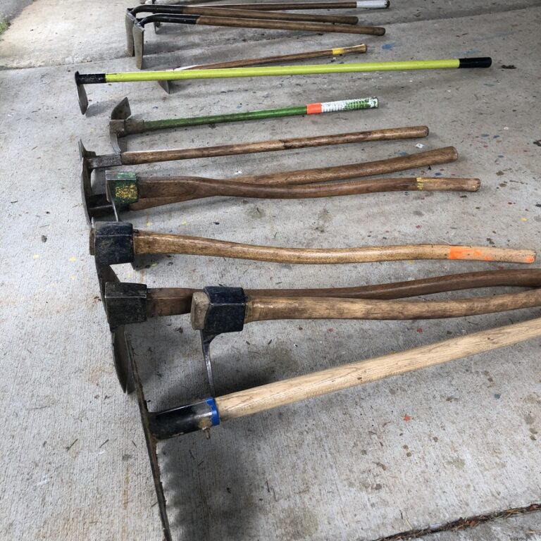 An assortment of had tools with long handles lay in a row on a concrete floor. 