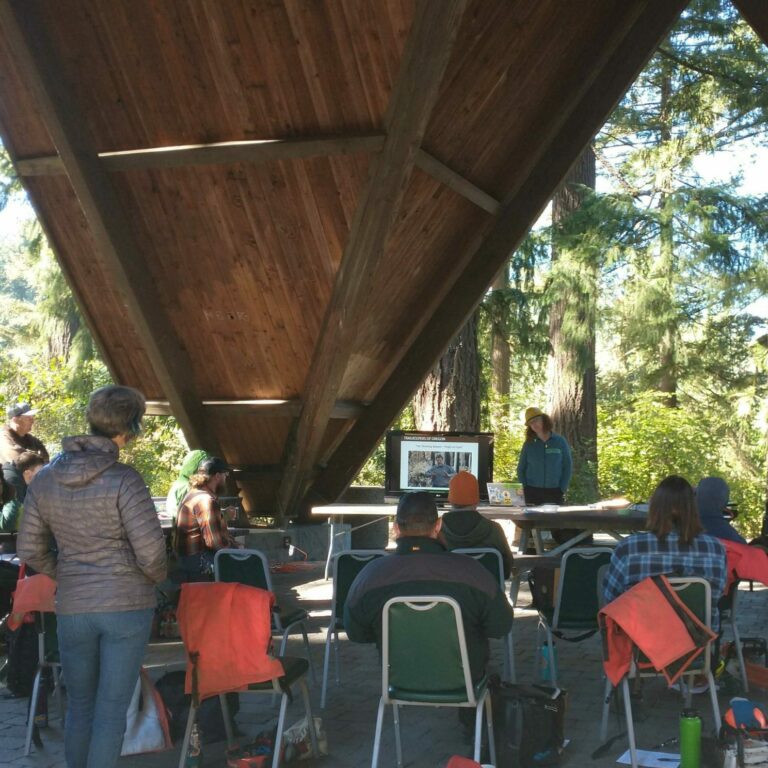 A group of people sit in chairs and watch a slideshow under a triangle shaped wooden shelter. 
