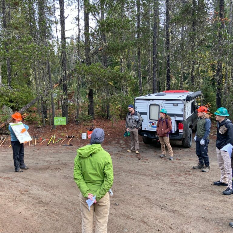 A group of people stand in a circle in a dirt parking lot. One person is using a large sign to teach the rest of the group about a trail maintenance topic. 