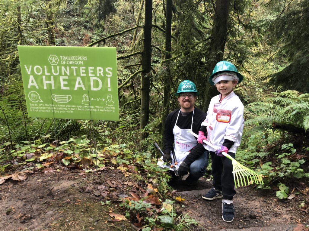 An adult and a child stand in the woods beside a green signs that says "Volunteers Ahead!". They are dressed as chefs, and the child holds a rake. 