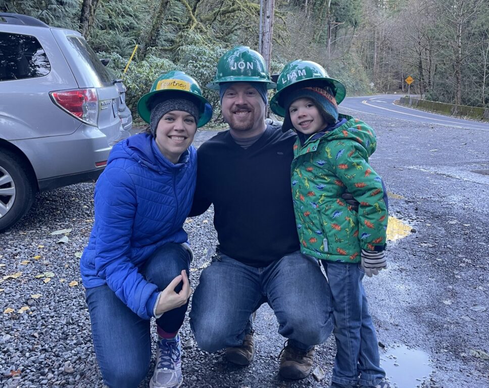 Two adults and a child, all wearing green hard hats, pose with their arms around each other while smiling at the camera. 