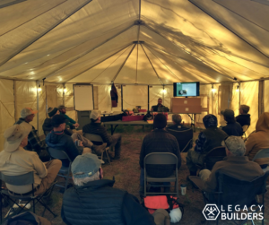 A group of volunteers sit inside a large tent listening to teachings from Saw Instructors.
