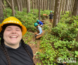 A TKO Crew Leader smiles taking a selfie while volunteers tend to the trail in the background.