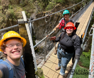 Three trail crew members pose smiling on a suspension bridge while holding a crosscut saw.