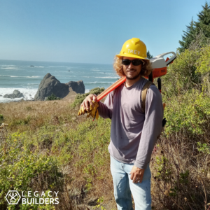 A person standing on a trail in a hardhat holds a chainsaw over their shoulder, the rocky Oregon coast in the background.