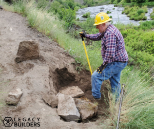 A volunteer in a yellow hardhat uses a long handled tool to move large rocks creating a hole in the trail surface.