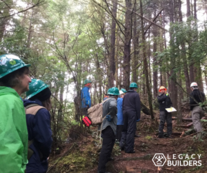 A group of volunteers stand on a forested trail listening to a Crew Leader.