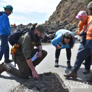 A group of TKO volunteers gather around a State parks Ranger who is describing a beach rock covered in small organisms.