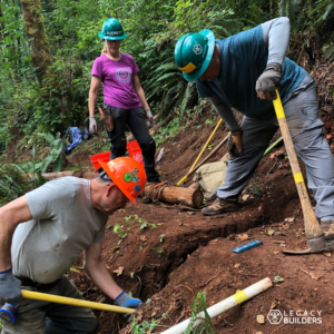 Three volunteers work together to dig a deep and narrow drainage ditch in the trail.