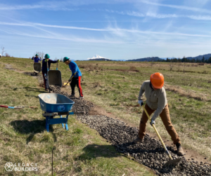 A group of volunteers spread gravel on a meadow trail with Mount Hood visible in the background.